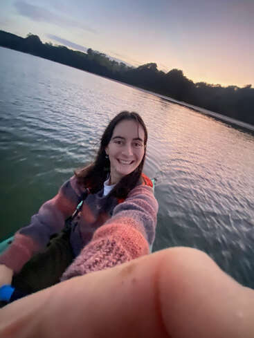Eine lächelnde Person macht ein Selfie beim Kajakfahren auf einem ruhigen See bei Sonnenuntergang. Der Himmel ist in sanften Farben gemalt und schafft eine friedliche Atmosphäre.