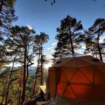 A imagem mostra uma tenda em forma de cúpula situada em uma área de floresta, com uma plataforma de madeira e uma vista serena do lago ao fundo em um dia ensolarado.