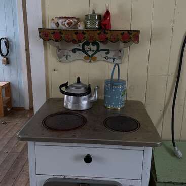 A imagem mostra um fogão de cozinha antigo com uma chaleira e um bule de chá em cima, tendo como pano de fundo uma parede e um piso de madeira.