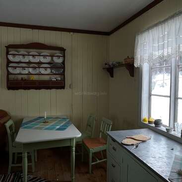 A imagem mostra uma área de jantar aconchegante com uma mesa, cadeiras e uma janela, com uma prateleira com pratos e uma bancada com uma tábua de corte e outros itens.
