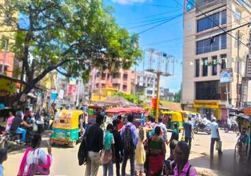 Scène de rue animée dans une ville indienne, avec des foules, des auto-rickshaws colorés, des motos, de grands bâtiments, des arbres, des vendeurs ambulants et un soleil radieux.