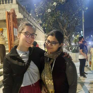 Two women pose for a photo on a city street at night, with a building, tree, and people in the background, under bright lights.