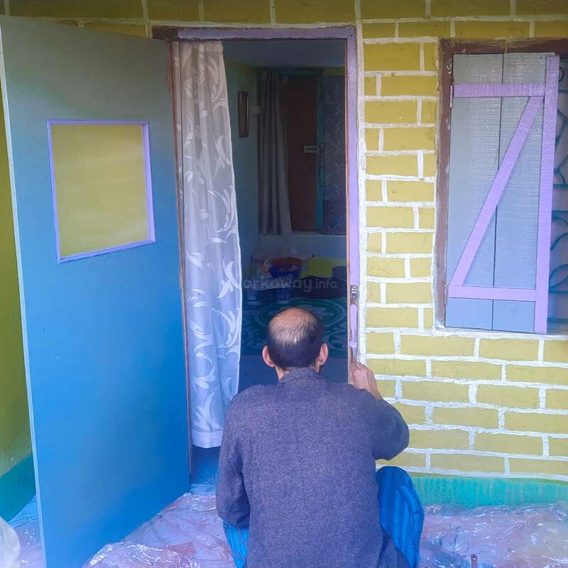 La imagen muestra a un hombre sentado frente a una casa con paredes de ladrillo amarillo, una puerta azul y una ventana, ocupado en una actividad.