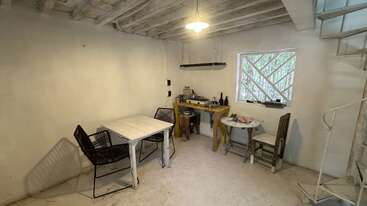 Cette pièce présente des murs et un plafond blancs de style rustique, une table simple avec des chaises, une petite table ronde, un établi en bois et un escalier en colimaçon.