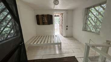 Cette chambre minimaliste est dotée d'un lit en bois blanc, d'une décoration rustique, de grandes fenêtres à barreaux et d'une porte donnant sur l'extérieur. La lumière naturelle inonde cet espace lumineux et épuré.