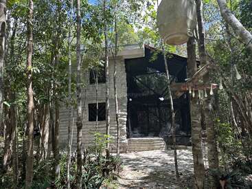 Une maison rustique en pierre nichée au cœur d'une forêt dense, entourée de grands arbres et de verdure. La lumière du soleil filtre à travers la canopée, créant une atmosphère sereine et paisible.
