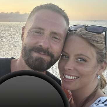 A happy couple poses for a selfie at the beach during sunset. They are smiling warmly, with the ocean and horizon visible in the background.