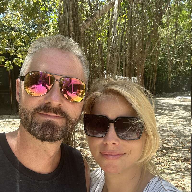 Un couple souriant, portant des lunettes de soleil, prend un selfie en plein air. La lumière du soleil filtre à travers les grands arbres en arrière-plan, créant une atmosphère chaleureuse et détendue.