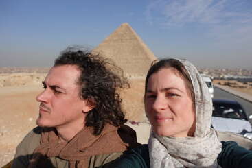 Un hombre y una mujer se hacen un selfi frente a la Gran Pirámide de Egipto, bajo un cielo azul y soleado. Ambos llevan bufandas y disfrutan de su aventura.