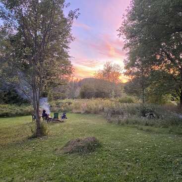 Una tranquila escena al aire libre muestra a dos personas sentadas junto a una hoguera, rodeadas de árboles y naturaleza, con una colorida puesta de sol que ilumina el cielo al fondo.