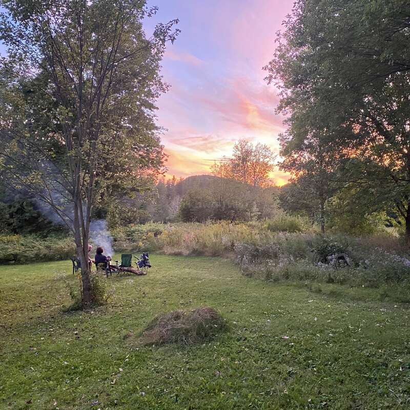 Una tranquila escena al aire libre muestra a dos personas sentadas junto a una hoguera, rodeadas de árboles y naturaleza, con una colorida puesta de sol que ilumina el cielo al fondo.