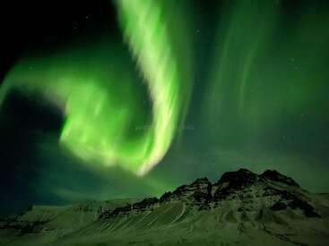 The image depicts the Northern Lights, also known as the Aurora Borealis, in a vibrant green hue, set against a dark sky and snow-capped mountains.