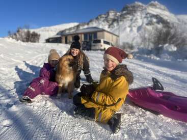 The image depicts three children and a dog sitting in the snow, with a house and mountains visible in the background on a sunny day.