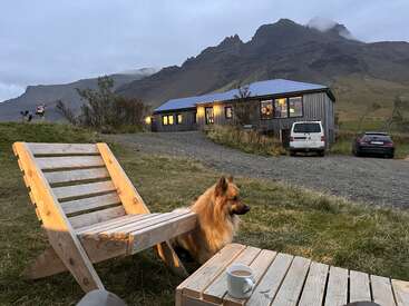 The image depicts a serene scene of a house, a dog, and a wooden chair with a coffee table, set against a mountainous backdrop on a cloudy day.