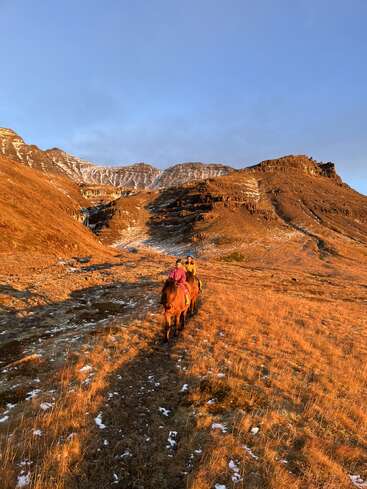 The image depicts two people riding horses on a trail in a mountainous, grassy area with patches of snow, set against a clear blue sky.