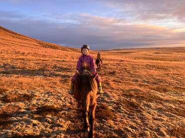 The image depicts two children riding horses through a vast, open field with rolling hills under a cloudy sky, likely during the golden hour.