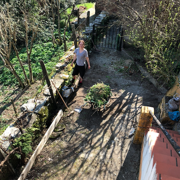 Eine Person steht draußen neben einer mit Gartenabfällen gefüllten Schubkarre, umgeben von Grün, Bäumen und Steinmauern – die Szene wird von oberhalb einer Treppe aus betrachtet.