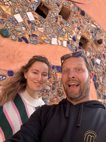 La imagen muestra a un hombre y una mujer tomándose un selfie delante de una pared cubierta de baldosas de cerámica rotas, con el hombre vestido con una sudadera negra con capucha y la mujer con un jersey de rayas.