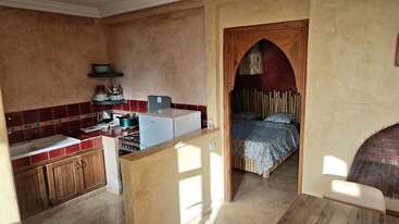 Esta imagen muestra una acogedora cocina con detalles en azulejos rojos, armarios de madera y una decoración rústica, situada junto a un dormitorio con una singular puerta en arco y un somier de madera.