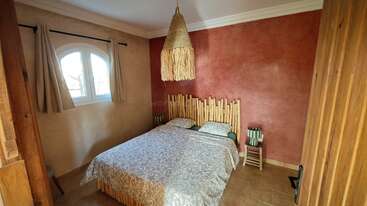 Este acogedor dormitorio cuenta con un cabecero rústico de madera, una cama doble con ropa de cama estampada, una lámpara colgante de mimbre, una ventana en arco, paredes con textura y una decoración sencilla y de tonos tierra.