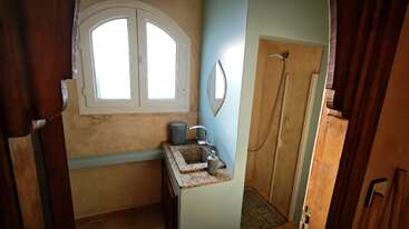 Esta imagen muestra un cuarto de baño pequeño con un lavabo rectangular de granito, una ventana en arco, detalles en azul claro, un espejo de pared y una zona de ducha abierta con una ducha de mano.