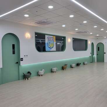 The image depicts a spacious room with white walls and green accents, featuring a row of animal-shaped stools and a large window with a school logo.