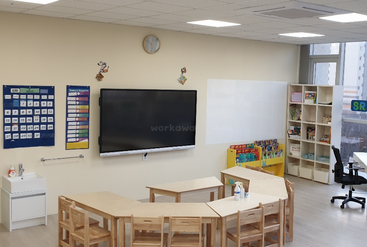 The image depicts a classroom with a table and chairs, featuring a whiteboard, a bookshelf, and a window. The room is well-lit, and the walls are painted white. The floor is made of wood.