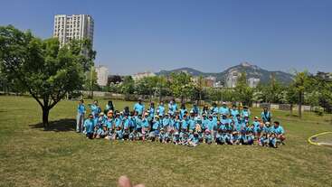The image shows a group of people gathered for a photo, with a large group of children in the foreground and a smaller group of adults in the background. The scene appears to be outdoors.