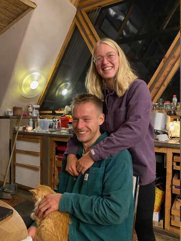 Um casal feliz sorri calorosamente em uma cozinha aconchegante. O homem, sentado, acaricia um gato laranja, enquanto a mulher fica atrás dele, com os braços carinhosamente em seus ombros.