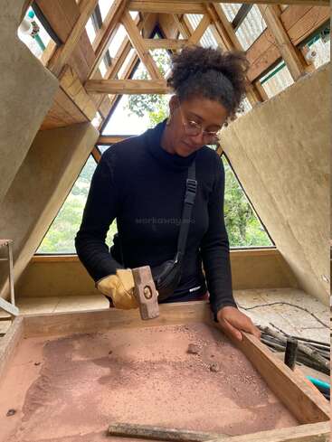 Uma mulher de óculos e blusa preta trabalha dentro de casa com luz natural, usando um martelo e uma luva para peneirar ou esmagar material em uma moldura de madeira.