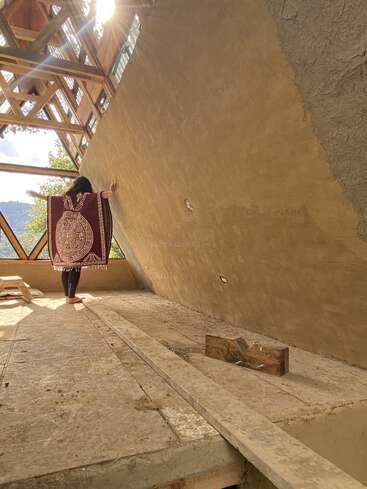 Uma pessoa está descalça em um piso de madeira dentro de um prédio ensolarado e parcialmente construído, com os braços estendidos, vestindo um poncho decorativo, com vigas de madeira angulares acima da cabeça.