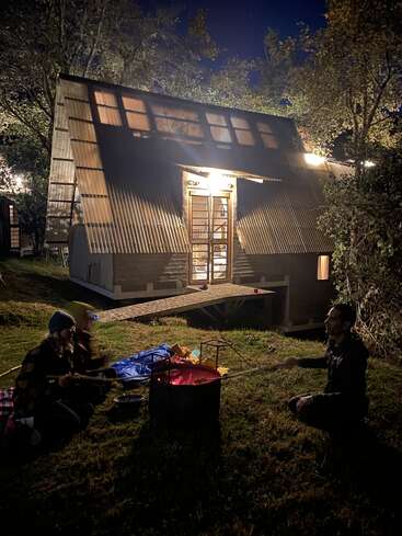 Duas pessoas sentam-se à noite ao lado de uma fogueira brilhante na grama, do lado de fora de uma cabana em forma de A bem iluminada e cercada por árvores, aproveitando uma noite tranquila ao ar livre.
