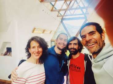 Quatro amigos sorridentes posam juntos dentro de casa, sob um teto de vidro brilhante e geométrico. O clima é aconchegante e convidativo, com luz natural e uma decoração confortável ao redor deles.