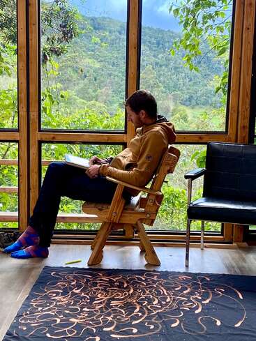 Um cara sentado dentro de casa numa cadeira de madeira, desenhando ou escrevendo num caderno, cercado por uma paisagem verdejante que dá pra ver pelas grandes janelas de vidro. Um clima tranquilo.