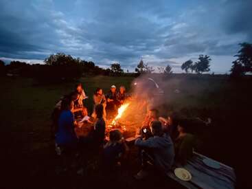 Eine Gruppe von Menschen versammelt sich in der Abenddämmerung um ein glühendes Lagerfeuer, um Geschichten und Wärme zu teilen, umgeben von Bäumen unter einem bewölkten, friedlichen Himmel in der Natur.