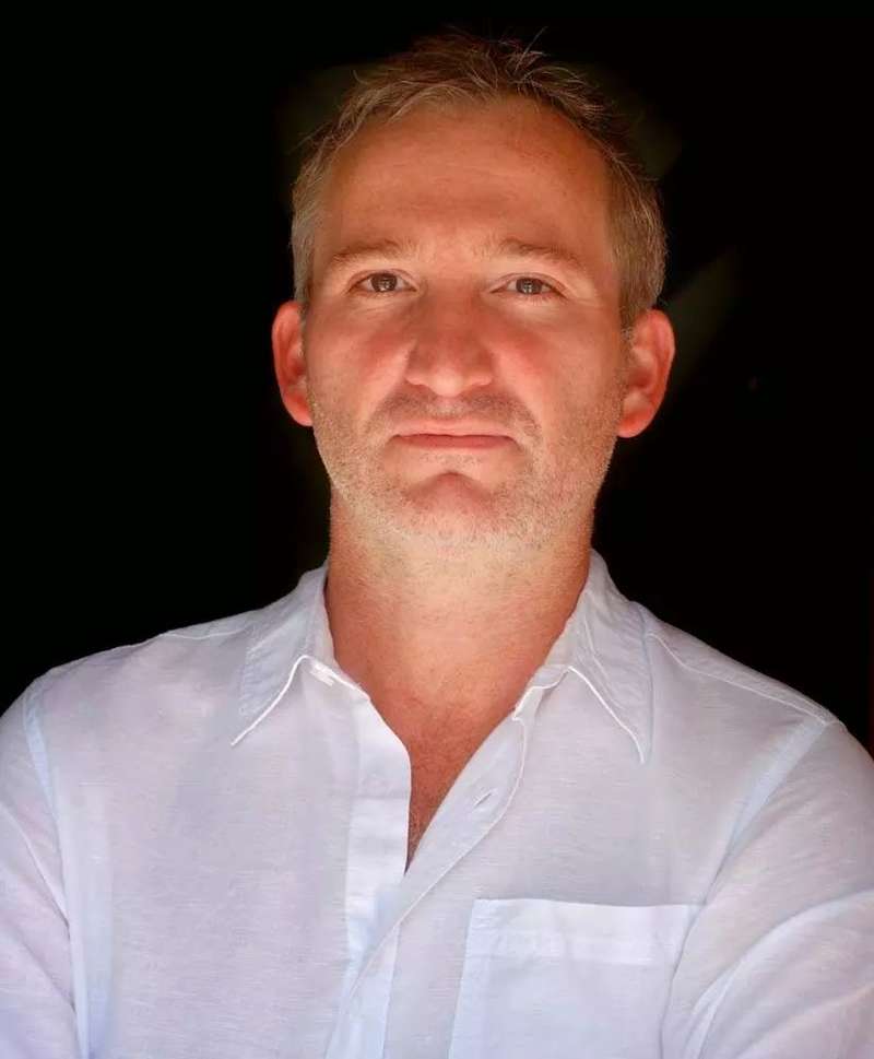 L'image représente un homme en chemise blanche, photographié à partir de la poitrine, avec une expression neutre et un sourire subtil, sur un fond noir austère.