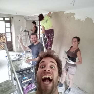 A imagem mostra um grupo de pessoas pintando uma sala, com uma pessoa tirando uma selfie em primeiro plano, capturando um momento de trabalho em equipe e criatividade.