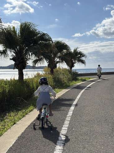 Uma criança anda de bicicleta com rodinhas atrás de um adulto numa estrada costeira ladeada por palmeiras, sob um céu claro e parcialmente nublado.