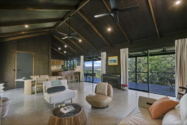 Ce salon spacieux et moderne est doté de hauts plafonds en bois, de grandes baies vitrées offrant une vue imprenable, d'un mobilier contemporain élégant, d'un coin repas, d'un piano, d'une cuisine et d'un accès à la terrasse extérieure.