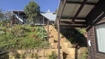 Une maison se dresse au sommet d'une colline, à laquelle on accède par un escalier en pierre entouré d'une végétation luxuriante, d'agaves et de parterres de fleurs, avec une terrasse couverte à l'avant.