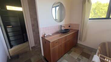 Cette salle de bains est équipée d'un meuble-lavabo en bois avec un lavabo noir, d'un miroir rond, de carreaux roses, d'une grande fenêtre avec des rideaux et d'un sol en pierre aux tons neutres. Ambiance chaleureuse.