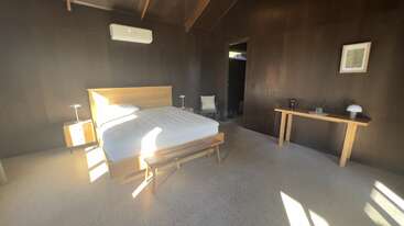 Cette chambre à coucher minimaliste comprend un lit en bois, deux tables de chevet, un éclairage tamisé, un bureau avec une lampe, des murs sombres, un sol blanc et bénéficie d'une abondante lumière naturelle.