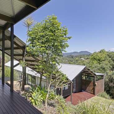 Une maison moderne au toit métallique nichée au cœur d'une végétation luxuriante, dotée d'une terrasse en bois et offrant une vue sur les montagnes sous un ciel bleu limpide. Un cadre paisible et ensoleillé.