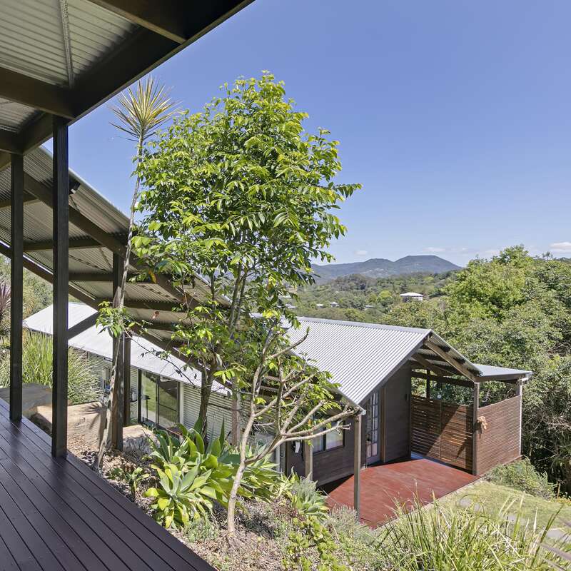 Une maison moderne au toit métallique nichée au cœur d'une végétation luxuriante, dotée d'une terrasse en bois et offrant une vue sur les montagnes sous un ciel bleu limpide. Un cadre paisible et ensoleillé.