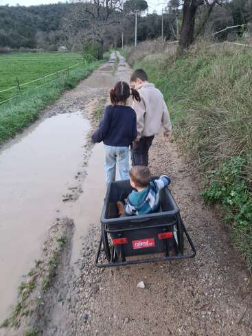 Drei Kinder laufen einen schlammigen Feldweg entlang; zwei gehen voraus, eines sitzt in einem schwarzen Wagen. Felder und Bäume umgeben ihr friedliches gemeinsames Abenteuer in der Natur.