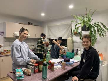Cuatro jóvenes adultos están en una acogedora cocina. Dos preparan la comida, uno friega los platos y otro está sentado y sonríe. Ingredientes y plantas decoran el espacio.
