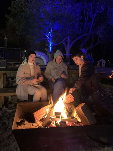 Tres personas se sientan alrededor de una hoguera encendida por la noche. Una persona añade leña. Al fondo se ven árboles iluminados de azul y un parque infantil que crean un ambiente acogedor.