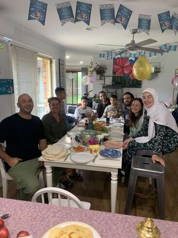 Una familia y sus amigos se reúnen alrededor de una mesa llena de comida para celebrar el Eid. La habitación está decorada con pancartas y globos de Eid Mubarak, creando un ambiente alegre.