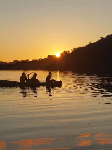 Three people paddle a canoe on a calm lake at sunset. The golden sky reflects beautifully on the water, creating a tranquil and peaceful scene.