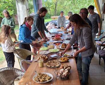 A group of people gathers around a long table filled with various dishes, enjoying a shared meal together in an outdoor, relaxed atmosphere surrounded by greenery.